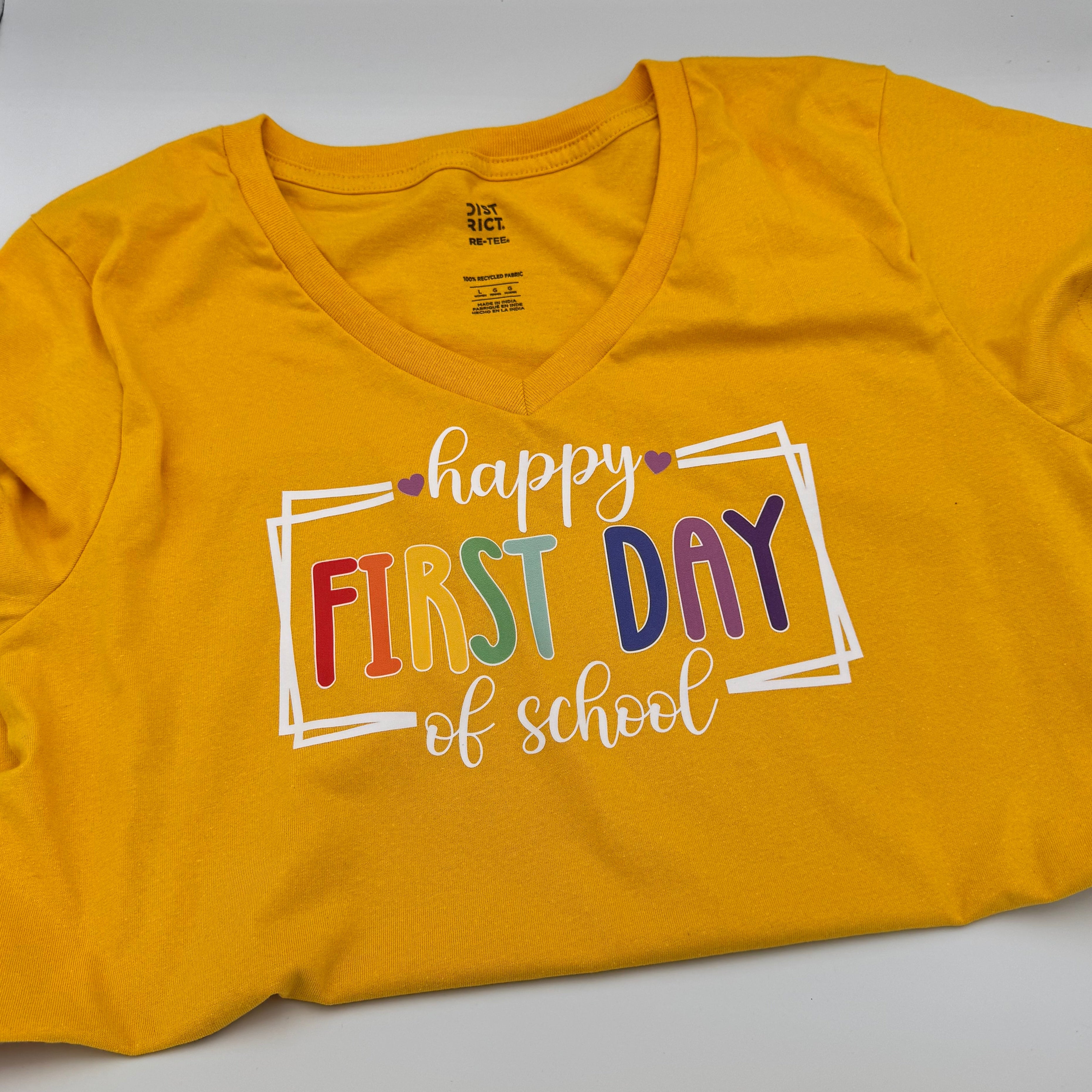 A yellow short-sleeve V-neck t-shirt laid flat on a white surface, featuring a colorful graphic that reads 'happy FIRST DAY of school' inside a white rectangular border.