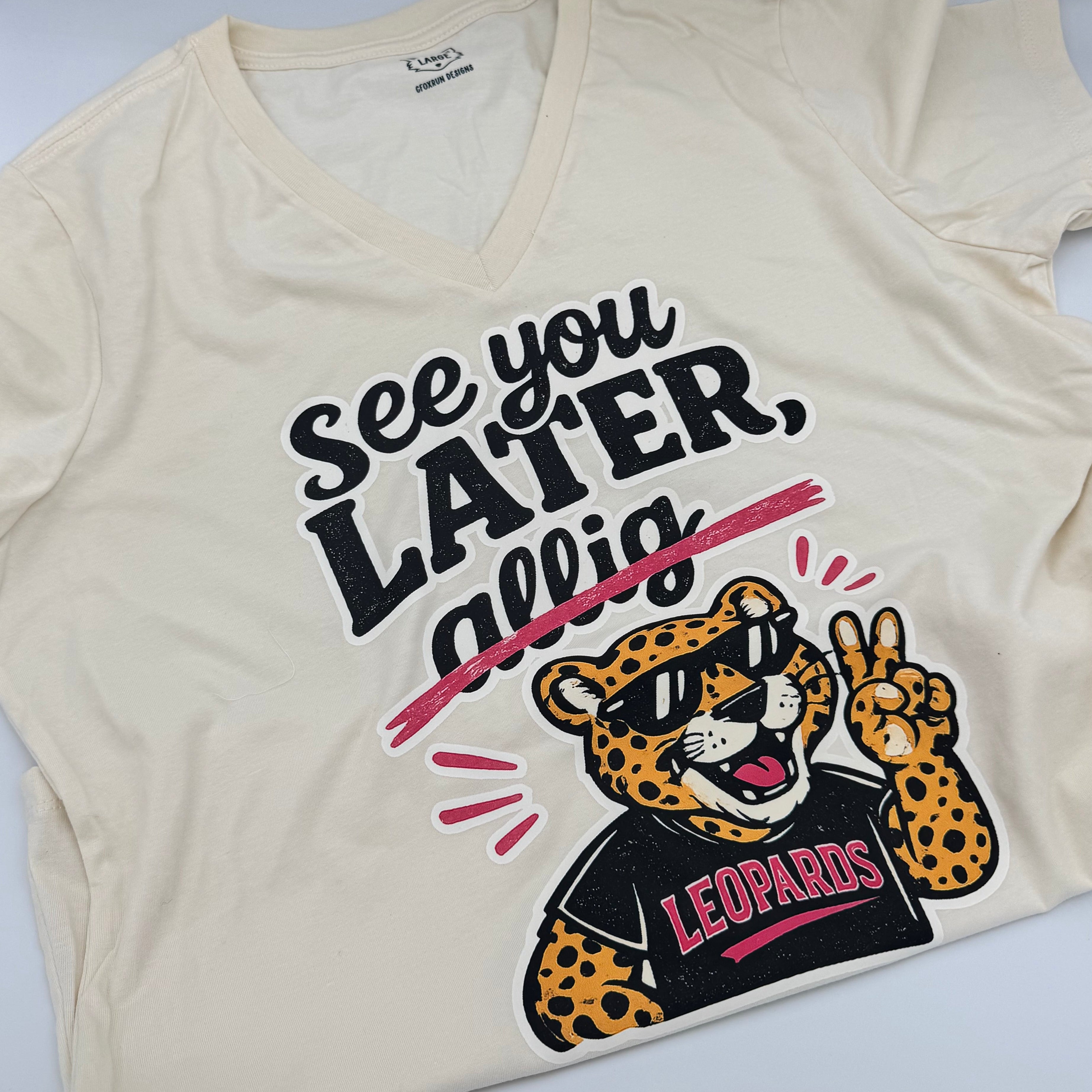 An off-white v-neck tee featuring a graphic of a leopard mascot wearing black sunglasses with 'LEOPARDS' written on it, alongside the text 'SEE YOU LATER, LEOPARDS!' in black and pink lettering.