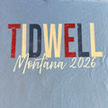 A light blue t-shirt laid flat, featuring a distressed graphic print that reads 'TIDWELL Montana 2026' in red, navy, and cream lettering.
