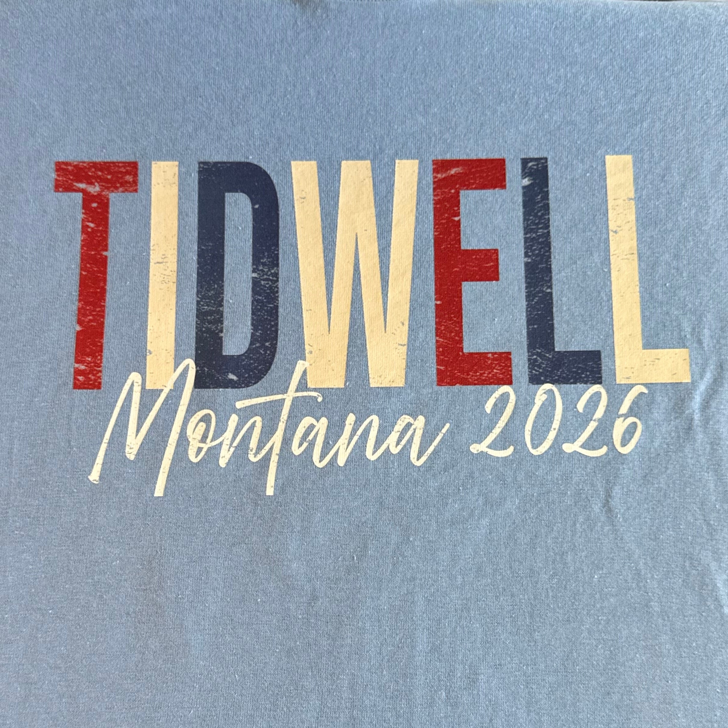 A light blue t-shirt laid flat, featuring a distressed graphic print that reads 'TIDWELL Montana 2026' in red, navy, and cream lettering.